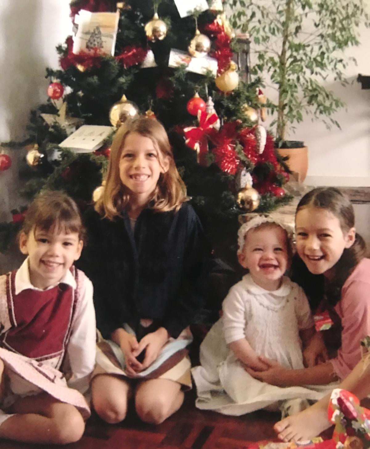 Four children sitting together in front of a decorated Christmas tree in a home setting, with holiday decorations, gifts, and a warm indoor atmosphere visible.